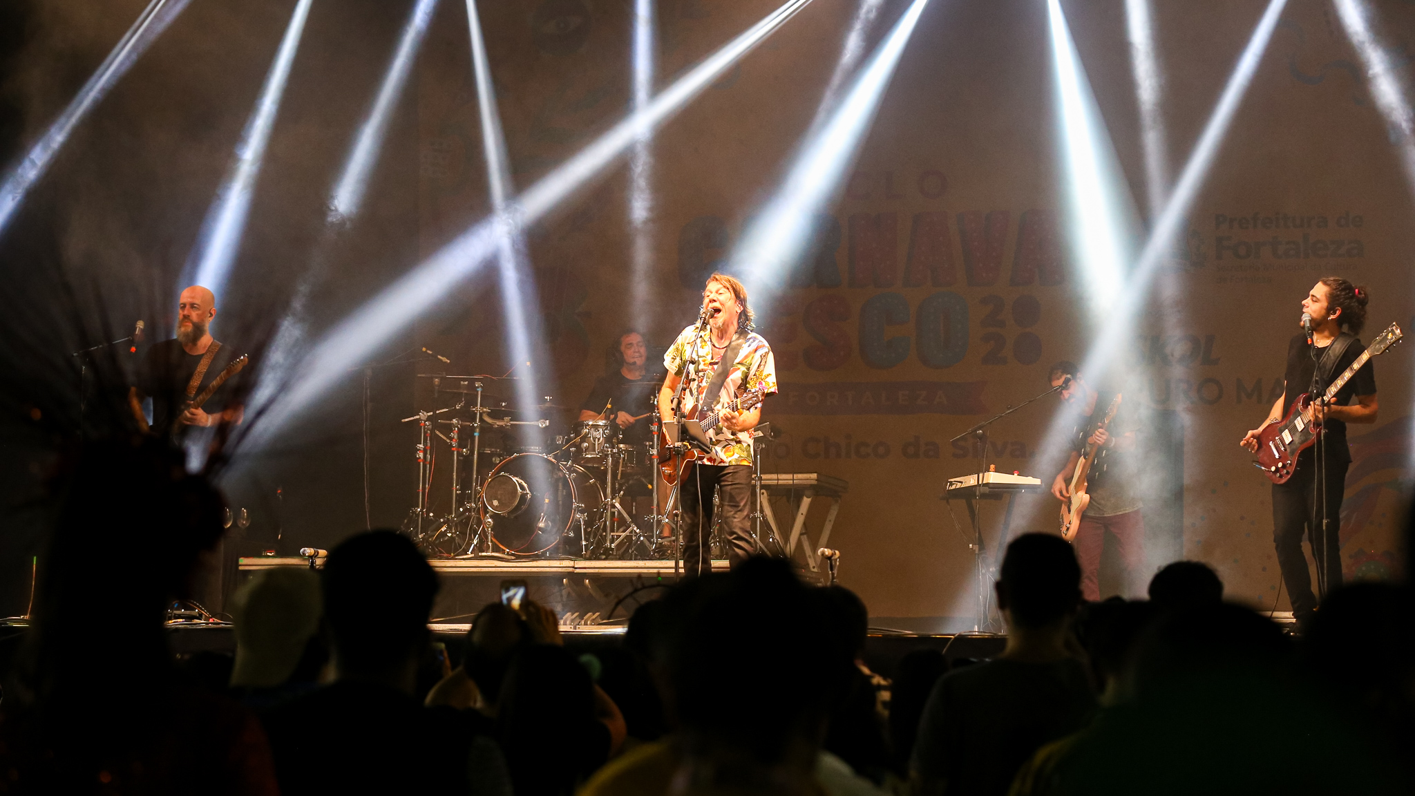 lenine tocando em palco ao lado de banda e pessoas em frente assistindo